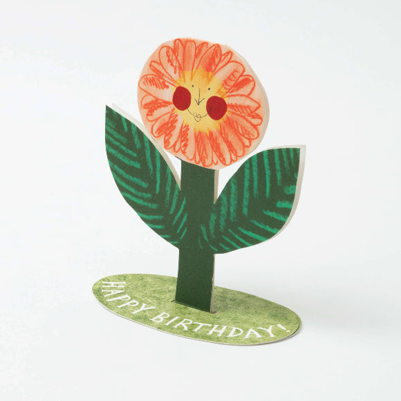 Aufstellkarte „Happy Birthday“ von Hadley: fröhliche Blumenfigur mit Gesicht, orange Blüte, grüne Blätter, ovaler Sockel mit „HAPPY BIRTHDAY!“.
