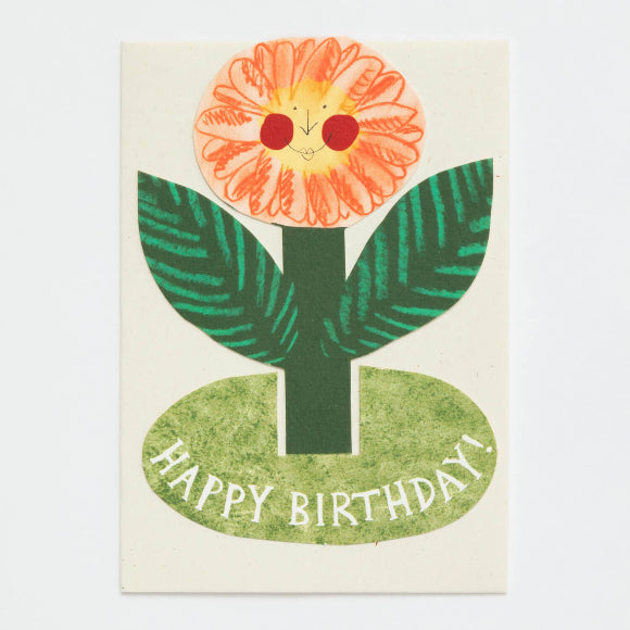 Aufstellkarte „Happy Birthday“ von Hadley: fröhliche Blumenfigur mit Gesicht, orange Blüte, grüne Blätter, ovaler Sockel mit „HAPPY BIRTHDAY!" und Umschlag.
